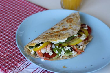 Karides, avokado, feta, ıspanak ve sebzelerle dolu katlanmış yumurtalı tortilla dürümü. Mavi tabakta servis edilmiş. Sağlıklı ve renkli kahvaltı konsepti