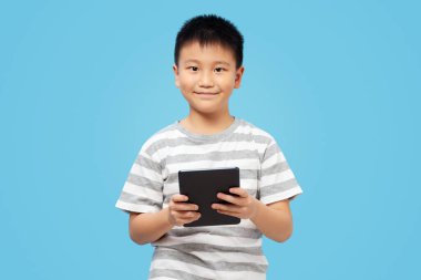 Akıllı çocuk elinde tabletle gülümsüyor ve kameraya bakıyor, mavi arka planda tişört giyiyor.