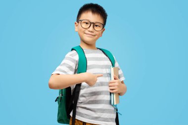 Sırt çantalı, gözlüklü, gülümseyen çocuk mavi arka plandaki kitapları işaret ediyor.