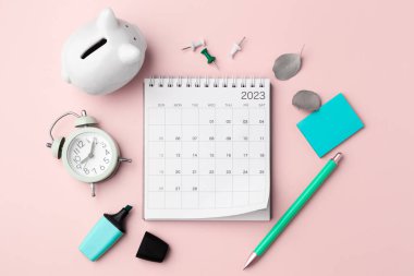 Calendar 2023 with stationary, clock and piggy bank on pastel pink background, top view