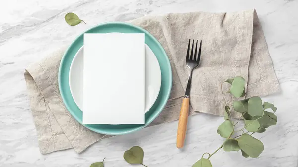 Wedding menu card on plate with fork on linen napkin on marble table with green leaves, top view