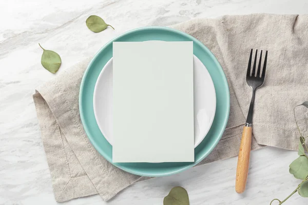 Green blank menu card mockup on plate with fork on linen napkin on marble table, top view