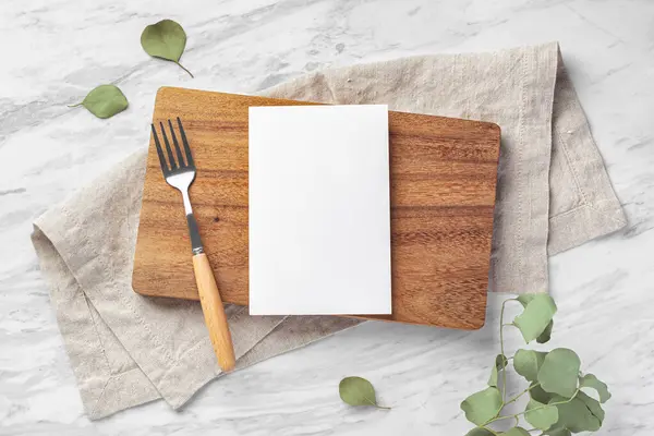 Wooden board and menu card mockup with fork and linen napkin on marble kitchen table with green leaves, top view