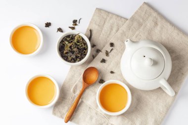 Keten kumaştan çay seti, kurutulmuş çay fincanları ve beyaz masa arkasında çaydanlık.