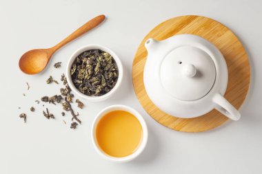 Bir fincan kurutulmuş çay, ahşap kaşık ve bej arka planda çaydanlık, üst manzara.