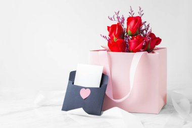 Gül buketli pembe hediye çantası ve siyah zarflı, kalbi beyaz kurdeleli bir kart.