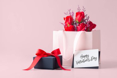 Kart, hediye kutusu ve pembe arka planda gül dolu sevgililer günün kutlu olsun.