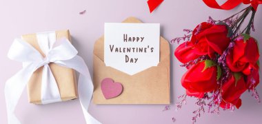 Kravat zarfında sevgililer günü kartı içinde kalp, hediye kutusu ve pembe arka planda gül buketi var.