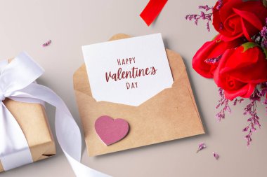 Kraft zarfında sevgililer günü kartı içinde kalp, hediye kutusu ve bej arka planda gül buketi var.