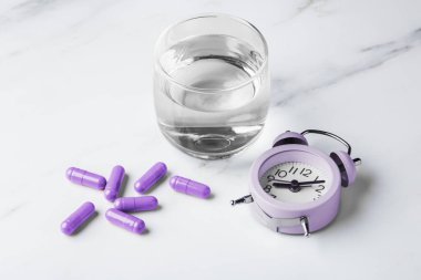 İlaç kapsülleri ve alarm saati olan bir bardak su, melatonin, takviye, beyaz masa arkasında vitamin konsepti