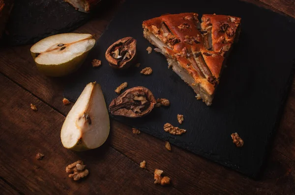 piece of pear pie on a black serving board on a wooden table, cut pears and walnuts lie chaotically