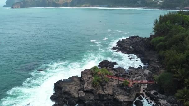 穏やかな海の波とサンゴの崖と緑の木と象徴的な赤い橋を持つビーチの美しさ ミュンチビーチ セントラルジャワ インドネシア — ストック動画