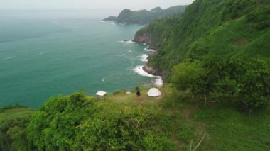 Bukit Jerit Kebumen 'in tepesinde kamp yapan bir adam tepeleri, yeşil ağaçları, mercanları ve okyanustan gelen dalgaları olan bir sahil şeridi.