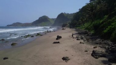 Drone View Pecaron Beach Kebumen 'de bir adam yürüyor, yeşil tepeli deniz kenarı, kum, mercan ve güzel dalgaları olan