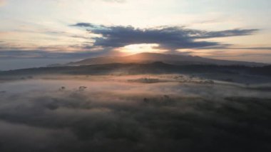 Kuş bakışı dramatik puslu gün doğumu. Güneş ışığı ağaçların arasından geçiyor. Tepeler ve arka planda bulutlar.