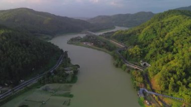 Serayu Nehri 'nin hava aracı görüntüsü, yan taraftaki karayolu ve demiryolu rayları, tepelerin arka planı, yeşil çam ormanları ve öğleden sonra parlak bulutlar.