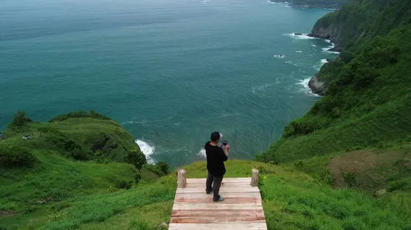 Bir adam deniz kenarında dalgalar, mercan resifleri ve kayalarla çevrili tepeler ve yeşil ağaçlarla bir platformda duruyor.