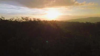 İnsansız hava aracı ağaçların ve ormanların üzerinden uçuyor. Günbatımı ve tepeler öğleden sonraya kadar arka planda kalıyor.