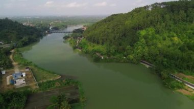Serayu Nehri 'nin hava aracı görüntüsü, yan taraftaki karayolu ve demiryolu rayları, tepelerin arka planı, yeşil çam ormanları ve öğleden sonra parlak bulutlar.