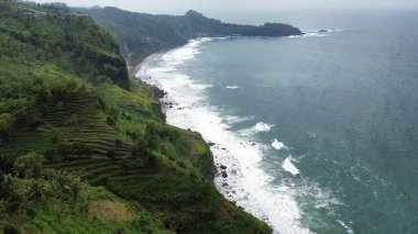 Tepeler ve ağaçlarla dolu kıyı şeridinin hava aracı görüntüsü, aynı zamanda mercan kayalıkları ve deniz manzarası Menganti Beach Kebumen Central Java Endonezya 'dan dalga dalga geliyor.