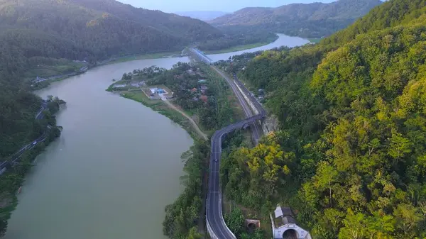 Serayu Nehri 'nin hava aracı görüntüsü, yan taraftaki karayolu ve demiryolu rayları, tepelerin arka planı, yeşil çam ormanları ve öğleden sonra parlak bulutlar.