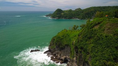 Tepeler ve ağaçlarla kıyı şeridini gösteren hava aracı manzarası, Watu Bale Beach Kebumen Central Java Endonezya 'daki mercan kayalıkları ve deniz dalgaları.
