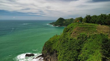 Tepeler ve ağaçlarla kıyı şeridini gösteren hava aracı manzarası, Watu Bale Beach Kebumen Central Java Endonezya 'daki mercan kayalıkları ve deniz dalgaları.