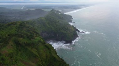 Tepeleri ve ağaçları olan kıyı şeridinin hava aracı görüntüsü, Sagara manzarasında okyanus dalgaları olan mercan kayalıkları ve deniz manzarası, Karang Bolong Beach Kebumen Merkezi Java Endonezya olarak da bilinir.