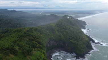 Tepeleri ve ağaçları olan kıyı şeridinin hava aracı görüntüsü, Sagara manzarasında okyanus dalgaları olan mercan kayalıkları ve deniz manzarası, Karang Bolong Beach Kebumen Merkezi Java Endonezya olarak da bilinir.