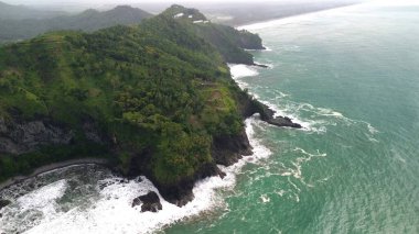 Tepeleri ve ağaçları olan kıyı şeridinin hava aracı görüntüsü, Sagara manzarasında okyanus dalgaları olan mercan kayalıkları ve deniz manzarası, Karang Bolong Beach Kebumen Merkezi Java Endonezya olarak da bilinir.
