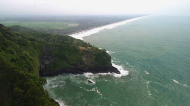 Tepeleri ve ağaçları olan kıyı şeridinin hava aracı görüntüsü, Sagara manzarasında okyanus dalgaları olan mercan kayalıkları ve deniz manzarası, Karang Bolong Beach Kebumen Merkezi Java Endonezya olarak da bilinir.