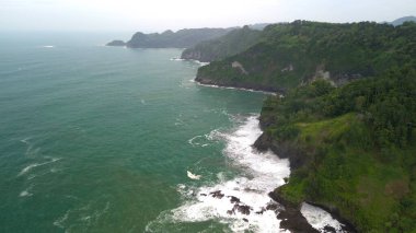 Tepeleri ve ağaçları olan kıyı şeridinin hava aracı görüntüsü, Sagara manzarasında okyanus dalgaları olan mercan kayalıkları ve deniz manzarası, Karang Bolong Beach Kebumen Merkezi Java Endonezya olarak da bilinir.