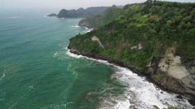 Tepeleri ve ağaçları olan kıyı şeridinin hava aracı görüntüsü, Sagara manzarasında okyanus dalgaları olan mercan kayalıkları ve deniz manzarası, Karang Bolong Beach Kebumen Merkezi Java Endonezya olarak da bilinir.