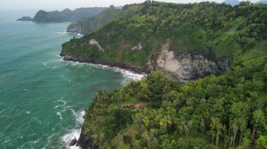 Tepeleri ve ağaçları olan kıyı şeridinin hava aracı görüntüsü, Sagara manzarasında okyanus dalgaları olan mercan kayalıkları ve deniz manzarası, Karang Bolong Beach Kebumen Merkezi Java Endonezya olarak da bilinir.