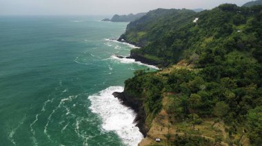 Tepeleri ve ağaçları olan kıyı şeridinin hava aracı görüntüsü, Sagara manzarasında okyanus dalgaları olan mercan kayalıkları ve deniz manzarası, Karang Bolong Beach Kebumen Merkezi Java Endonezya olarak da bilinir.