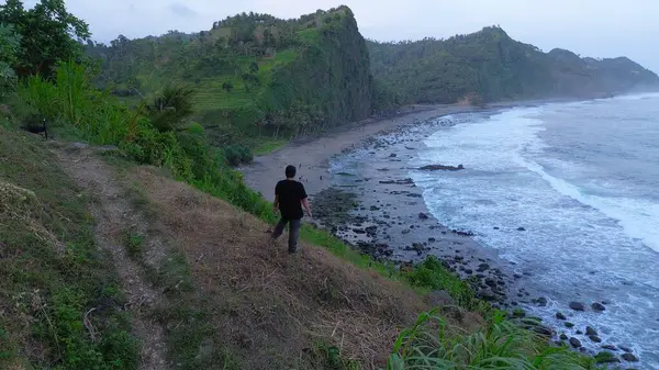  Tepelerin, kayalıkların, ağaçların, kumsalların, kumsalların ve dalgaların bulunduğu bir adam Karang Bolong Sahili, Kebumen, Central Java, Endonezya