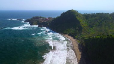 Tepeler ve ağaçlarla dolu kıyı şeridinin hava aracı görüntüsü, mercan kayalıkları ve deniz manzarası, Pecaron Beach Kebumen Central Java Endonezya 'daki okyanus dalgaları.