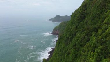 İnsansız hava aracı manzaralı seyahat rotası, tepeli ve ağaçlı kıyı şeridi, okyanustan gelen dalgalar mercan resiflerine çarpıyor, Jerit Hill ve Beach Kebumen Central Java Endonezya
