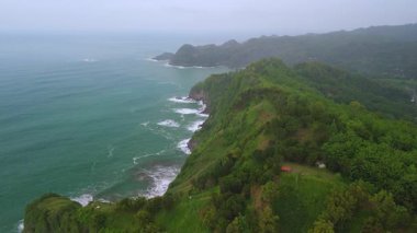 İnsansız hava aracı manzaralı seyahat rotası, tepeli ve ağaçlı kıyı şeridi, okyanustan gelen dalgalar mercan resiflerine çarpıyor, Jerit Hill ve Beach Kebumen Central Java Endonezya