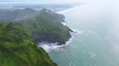 İnsansız hava aracı manzaralı seyahat rotası, tepeli ve ağaçlı kıyı şeridi, okyanustan gelen dalgalar mercan resiflerine çarpıyor, Sagara manzaralı sahil Kebumen Central Java Endonezya