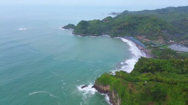 İnsansız hava aracı manzaralı seyahat rotası, tepeli ve ağaçlı kıyı şeridi, okyanustan gelen dalgalar mercan resiflerine çarpıyor, Watu Bale Beach Kebumen Central Java Endonezya