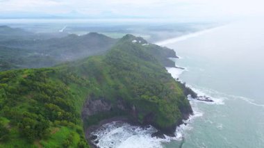 İnsansız hava aracı manzaralı seyahat rotası, tepeli ve ağaçlı kıyı şeridi, okyanustan gelen dalgalar mercan resiflerine çarpıyor, Jerit Hill ve Beach Kebumen Central Java Endonezya