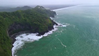 İnsansız hava aracı manzaralı seyahat rotası, tepeli ve ağaçlı kıyı şeridi, okyanustan gelen dalgalar mercan resiflerine çarpıyor, Sagara manzaralı sahil Kebumen Central Java Endonezya