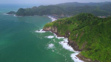 İnsansız hava aracı manzaralı seyahat rotası, tepeli ve ağaçlı kıyı şeridi, okyanustan gelen dalgalar mercan resiflerine çarpıyor, Watu Bale Beach Kebumen Central Java Endonezya