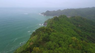İnsansız hava aracı manzaralı seyahat rotası, tepeli ve ağaçlı kıyı şeridi, okyanustan gelen dalgalar mercan resiflerine çarpıyor, Jerit Hill ve Beach Kebumen Central Java Endonezya