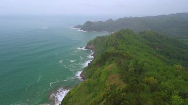 İnsansız hava aracı manzaralı seyahat rotası, tepeli ve ağaçlı kıyı şeridi, okyanustan gelen dalgalar mercan resiflerine çarpıyor, Jerit Hill ve Beach Kebumen Central Java Endonezya