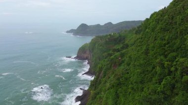İnsansız hava aracı manzaralı seyahat rotası, tepeli ve ağaçlı kıyı şeridi, okyanustan gelen dalgalar mercan resiflerine çarpıyor, Jerit Hill ve Beach Kebumen Central Java Endonezya