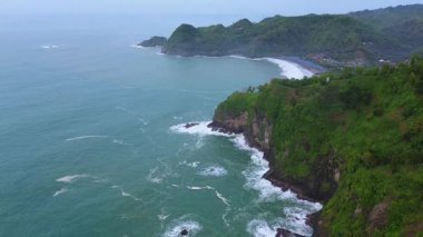 İnsansız hava aracı manzaralı seyahat rotası, tepeli ve ağaçlı kıyı şeridi, okyanustan gelen dalgalar mercan resiflerine çarpıyor, Watu Bale Beach Kebumen Central Java Endonezya