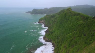 İnsansız hava aracı manzaralı seyahat rotası, tepeli ve ağaçlı kıyı şeridi, okyanustan gelen dalgalar mercan resiflerine çarpıyor, Watu Bale Beach Kebumen Central Java Endonezya