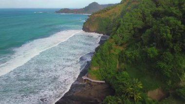Öğleden sonra Surumanis, Kebumen, Central Java, Endonezya 'da kıyı şeridinde okyanus manzaralı ve mercan kayalıklarına vuran dalgalarla bir tepe ve sahil insansız hava aracı.